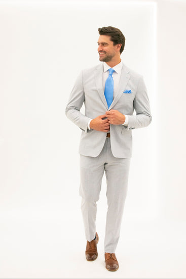 Man wearing a silver gray suit with a blue tie and pocket square on a white background looking to the left while smiling and holding the buttons of the jacket. 