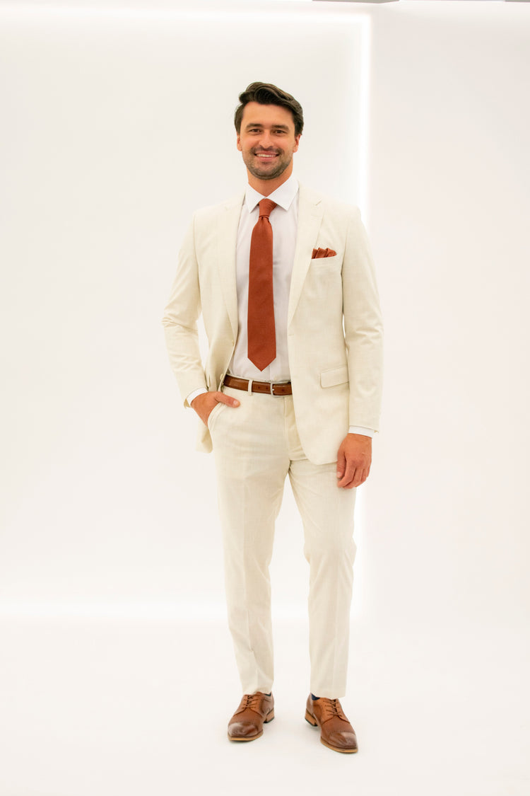 Man wearing a tan suit jacket unbuttoned with tan suit trousers along with a rust colored tie and pocket square looking to the right. Accessorized with a white dress shirt, cognac belt, and cognac shoes with his hand in his pant pocket. Grooms Club. 