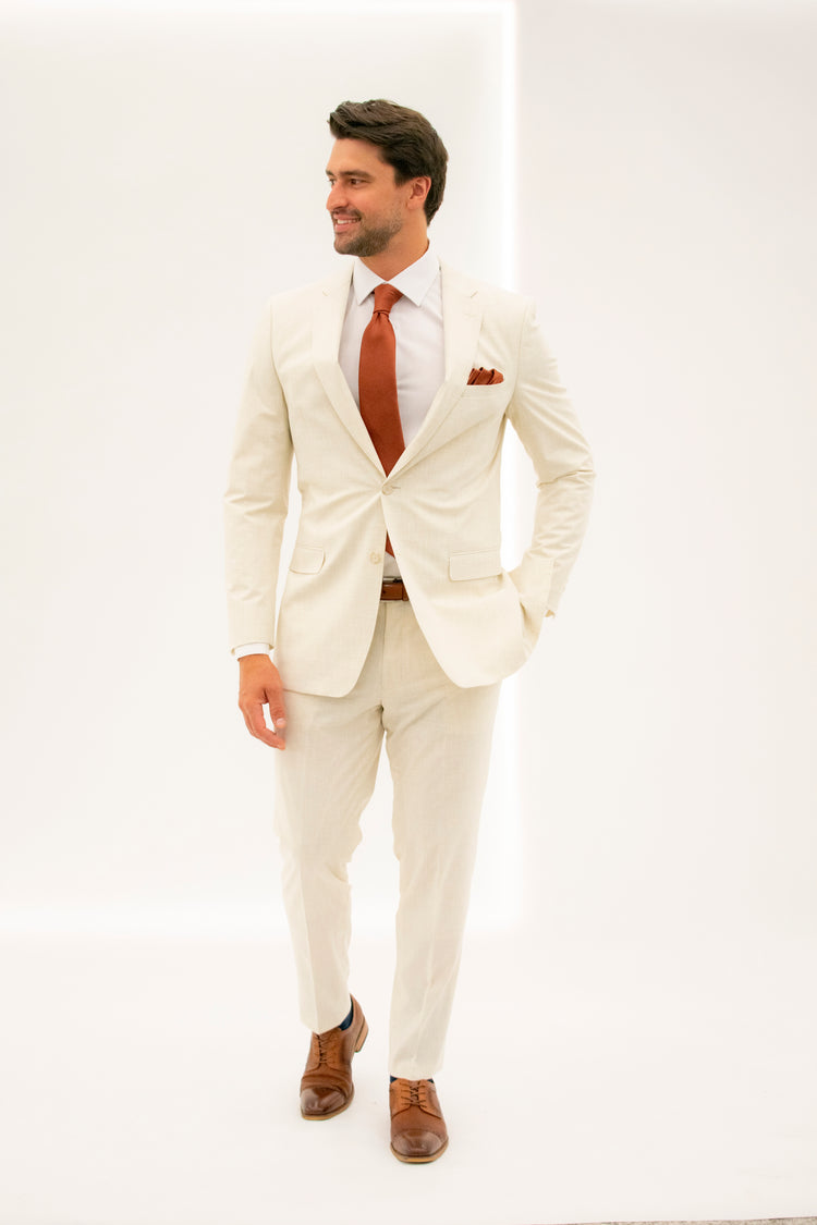 Man wearing a tan suit jacket with tan suit trousers along with a rust colored tie, pocket square, cognac shoes, and a cognac belt looking to the left. Grooms Club. 