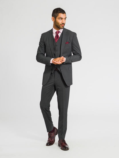 A man wearing a charcoal gray jacket, charcoal gray vest, and charcoal gray pants with a burgundy tie, burgundy pocket square, and burgundy shoes against a white background looking off to the right. 