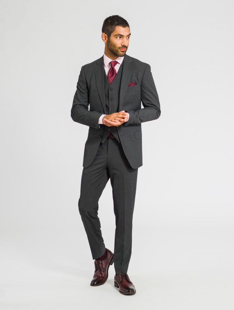 A man wearing a charcoal gray jacket, charcoal gray vest, and charcoal gray pants with a burgundy tie, burgundy pocket square, and burgundy shoes against a white background looking off to the right. 