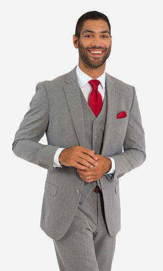 A man wearing a light grey suit jacket, light grey vest, and light grey pants with a red tie and a matching handkerchief, standing against a white background smiling. 