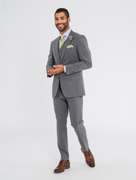 Man facing camera smiling wearing a three piece medium grey jacket, medium grey vest, medium grey pants, a sage tie, sage pocket square, sage lapel pin, and cognac shoes. Grooms Club. 