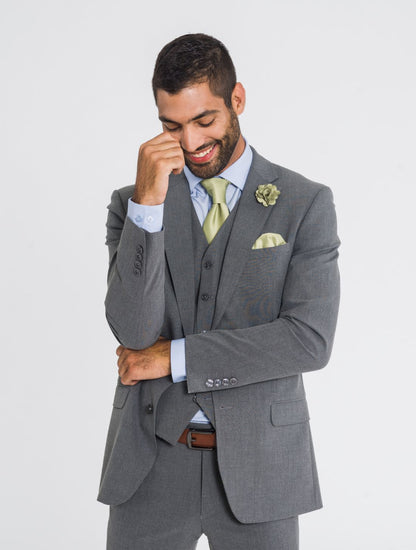 Close up image of a man wearing a three piece medium grey jacket, medium grey vest, medium grey pants, a sage tie, sage pocket square, sage lapel pin, and a cognac belt while smiling and looking down. Grooms Club. 