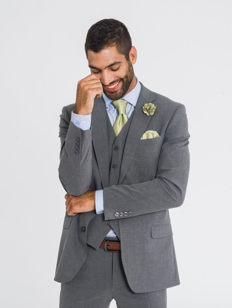 Close up image of a man wearing a three piece medium grey jacket, medium grey vest, medium grey pants, a sage tie, sage pocket square, sage lapel pin, and a cognac belt while smiling and looking down. Grooms Club. 