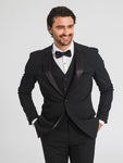 A zoomed in image of man wearing a black tuxedo jacket, a black tuxedo vest, black tuxedo pants, a white tuxedo shirt, and a black bowtie smiling while looking to the right past the camera. 
