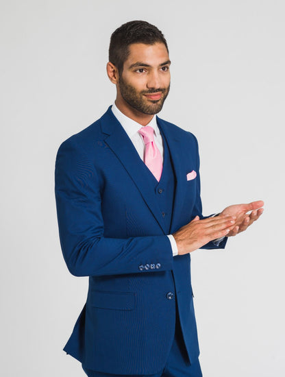 Close up image of  man wearing a blue suit with a jacket, vest, trousers, and a pink tie and pocket square standing against a white background - Grooms Club. 