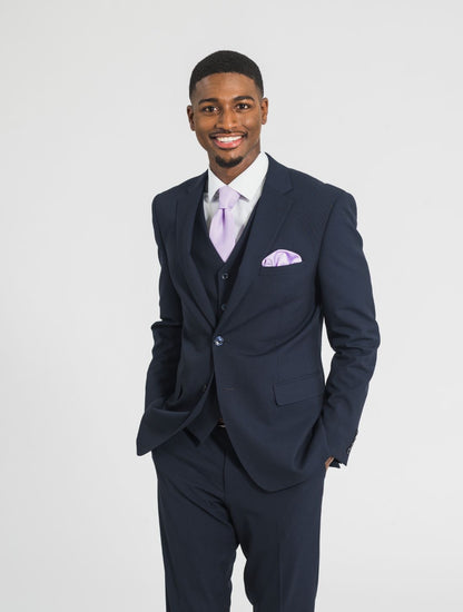 Close up image of man wearing a navy jacket, navy vest, and navy pants, with a lavender tie and lavender pocket square, against a white background smiling. Grooms Club. 