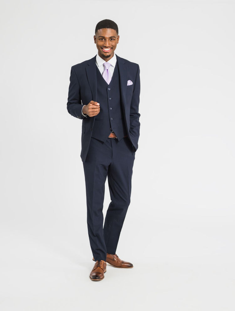 Zoomed out image of a man wearing a three piece navy suit ensemble along with brown shoes, a brown belt, a lavender tie, and a lavender pocket square for a wedding. Grooms Club.  