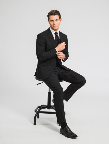 A zoomed out image of a man sitting on a chair wearing a black suit jacket, a black vest, black pants, along with a black belt,  black shoes, a black tie, and a black pocket square leaning to the side smiling at the camera. 