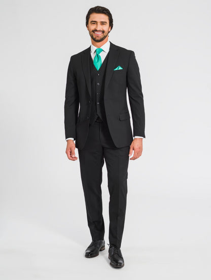 A zoomed out image of a man wearing a black suit jacket, a black vest, black pants, along with a black belt,  black shoes, a teal tie, and a teal pocket square facing forward smiling at the camera. 