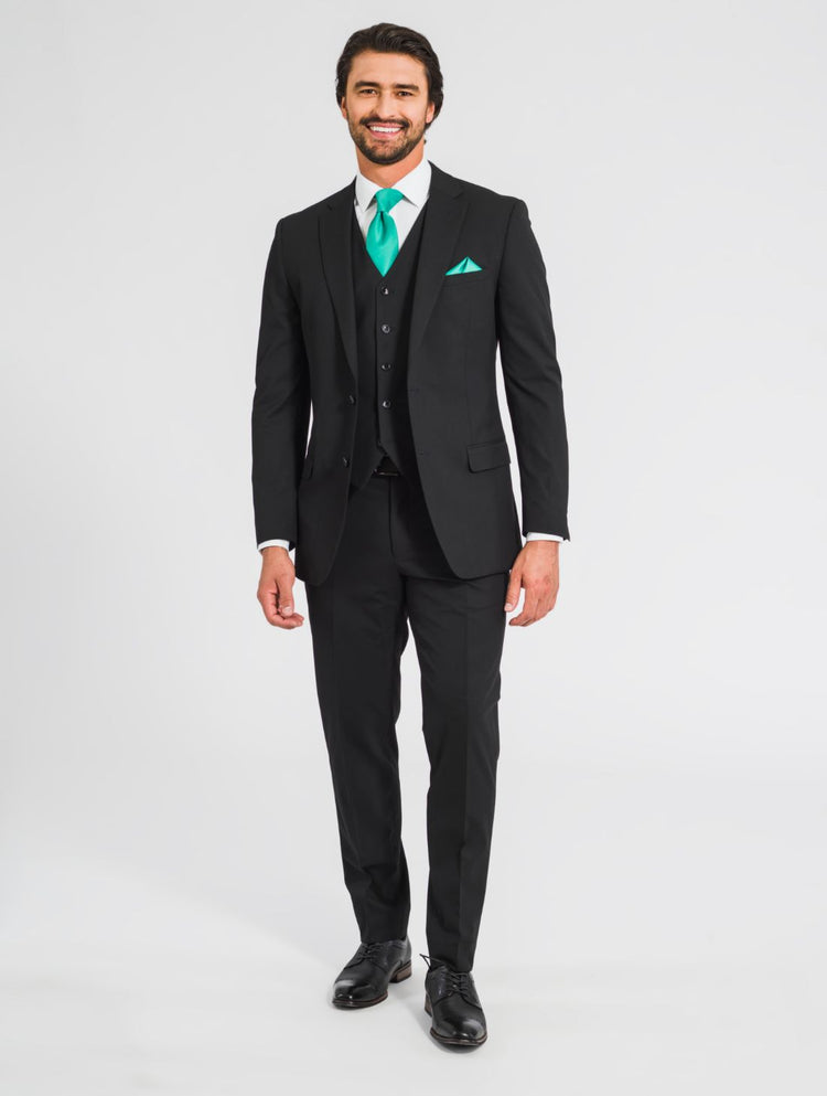 A zoomed out image of a man wearing a black suit jacket, a black vest, black pants, along with a black belt,  black shoes, a teal tie, and a teal pocket square facing forward smiling at the camera. 